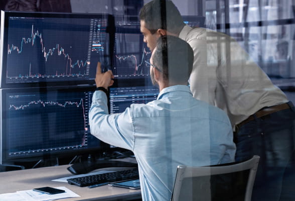 Two men analyzing stock market data on computer screens, staying informed about foreign exchange and interest rate trends for effective risk management